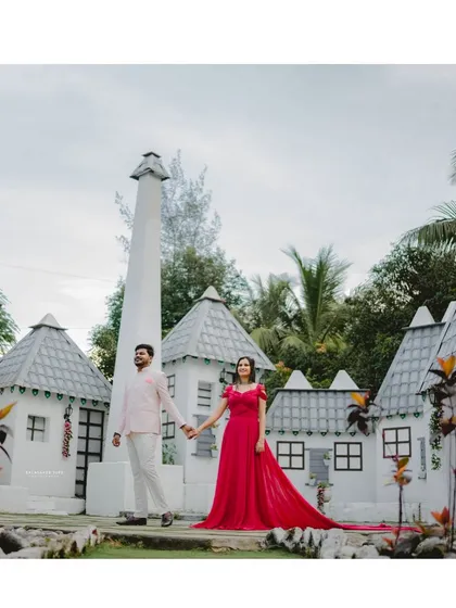 A wide shot showing the full splendor of the magenta trail gown against a whimsical, village-like backdrop for a unique pre-wedding photo.