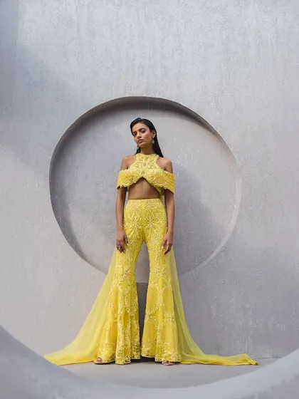 This stunning sunshine yellow sharara set is a masterpiece of intricate embellishments, with a cold-shoulder silhouette and an attached floor-sweeping drape.
