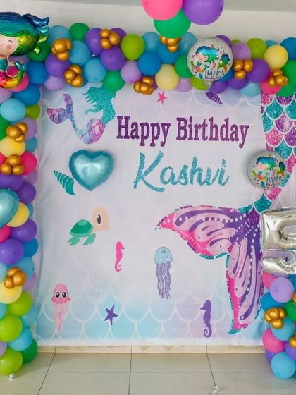 A colorful mermaid-themed birthday decoration for a 5th birthday. The setup includes a custom backdrop, a vibrant balloon frame, and mermaid and sea creature foil balloons.