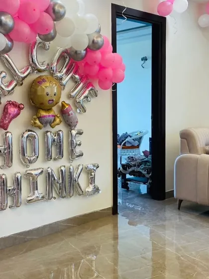 A closer look at a personalized "Welcome Home" wall decoration. The silver foil balloons are accented with pink and silver balloon clusters and transparent 'BABY' blocks filled with pink balloons.
