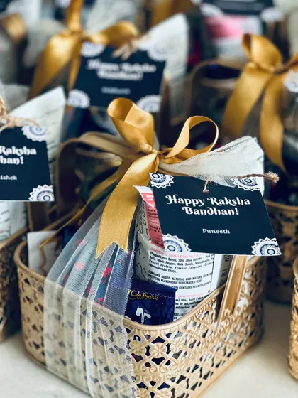 We design thoughtful gifts for every festival, like these elegant Raksha Bandhan hampers. Each golden metal basket is filled with treats and a travel mug, finished with a personalized "Happy Raksha Bandhan" tag for a special touch.