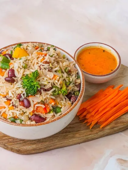 A healthy vegetable and kidney bean rice bowl, served on a wooden platter with a side of tangy sauce and julienned carrots. The light, airy background makes this dish look fresh and appealing for a healthy menu.