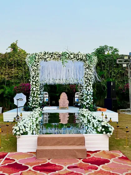 A stunning wedding ceremony stage with a mirrored aisle reflecting the grand floral arch. The white floral arrangements and hanging tassels create a magical and ethereal atmosphere.