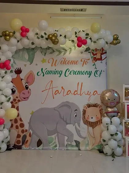 A complete jungle-themed naming ceremony decoration. This setup includes a custom backdrop with cartoon animals, a white and gold balloon arch, and "BABY" letter blocks, creating a perfect spot for photos.
