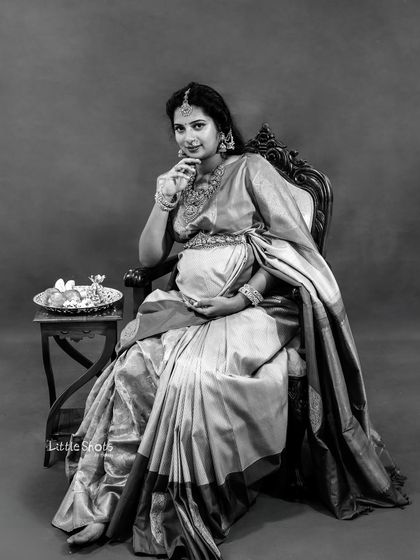 A classic black and white portrait of a mother-to-be seated in a traditional saree. She looks directly at the camera, her pose and expression creating a timeless and elegant photograph.