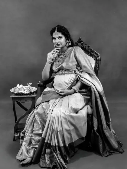 A classic black and white portrait of a mother-to-be seated in a traditional saree. She looks directly at the camera, her pose and expression creating a timeless and elegant photograph.