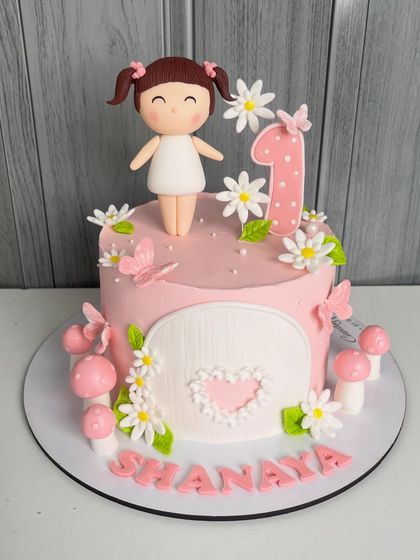 A full view of the baby pink first birthday cake, personalized with the name "Shanaya." The design features a charming doll topper, butterflies, and a whimsical mushroom house theme.