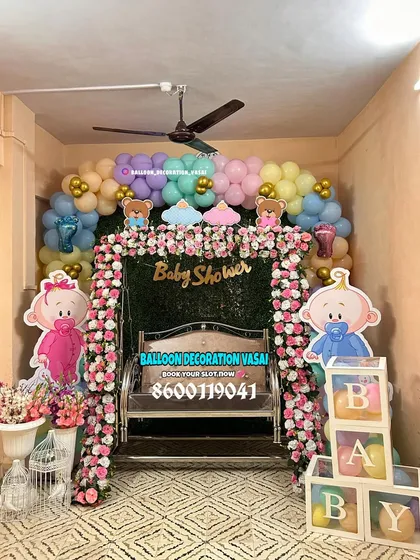 This cozy baby shower setup features a lush green wall backdrop framed with flowers and pastel balloons. The decorated jhula and "BABY" blocks complete this charming arrangement for a home celebration.