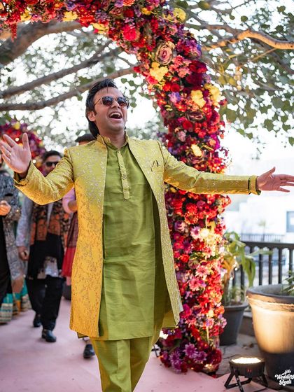 The groom makes a grand, joyful entrance under a floral arch, his arms wide open to embrace the celebration.