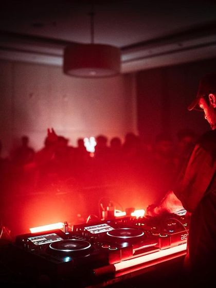 A DJ seen from behind the decks, looking out over the crowd. The red lighting and haze create a powerful, immersive atmosphere that defines our techno nights.