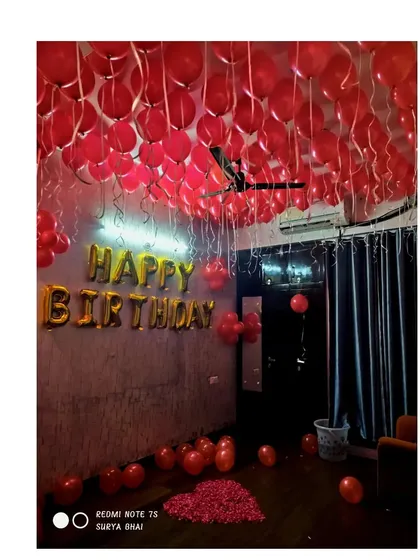 A simple and classic birthday setup in a room with low lighting. The ceiling is covered with red balloons, and 'Happy Birthday' foil letters are the main focus on the wall.