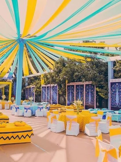 A large-scale Mehendi setup with a canopy of yellow and blue drapes. The decor includes both low floor seating and tables with chairs to accommodate all guests comfortably.