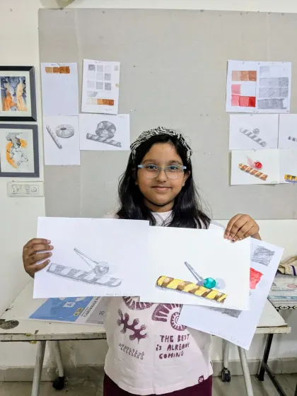 This student proudly presents her completed object study. She has created both a pencil sketch and a colored version, demonstrating her understanding of shading and color.