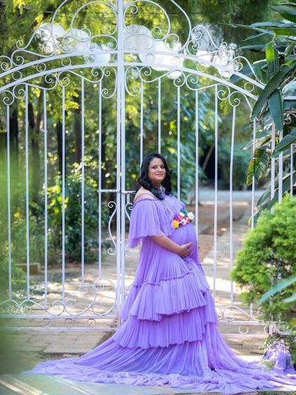 A lovely portrait of a mom-to-be in a lavender gown and floral belt, standing by an elegant white gate.