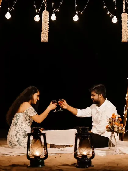 A couple toasts under a canopy of fairy lights on the beach at night. This image showcases a beautifully styled, romantic picnic setup perfect for a pre-wedding shoot.