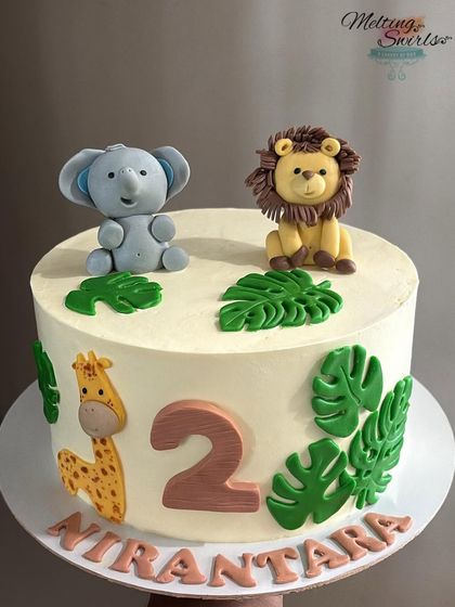 A simple and cute jungle-themed cake perfect for a second birthday. This design features adorable, handmade fondant animals like an elephant, lion, and giraffe on a clean, white whipped cream base.
