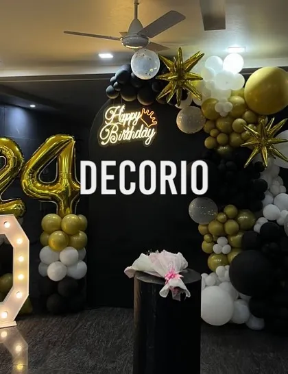 A stylish 24th birthday decoration with a black, gold, and white color scheme. The setup includes a large 'D' marquee letter, '24' foil balloons, and a balloon arch against a black backdrop with a neon sign.