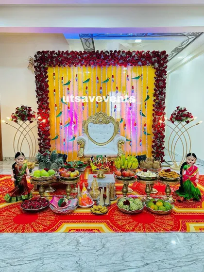 A beautiful baby shower stage with a yellow backdrop framed by red roses. The setup includes a white throne and a full display of traditional offerings on a red carpet.