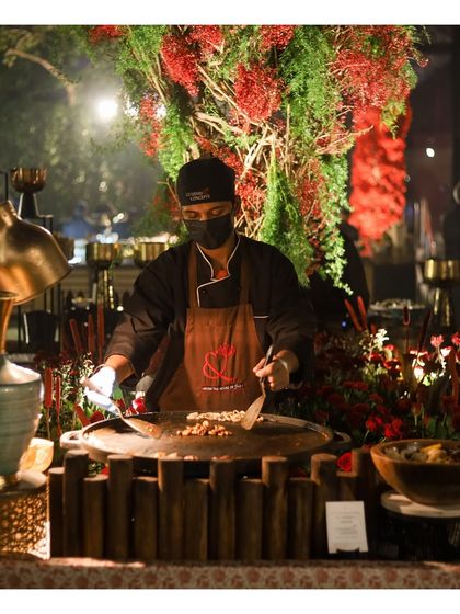 A chef stir fries noodles or vegetables on a large wok at an evening event. The dramatic lighting and lush decor create a sophisticated atmosphere for our live cooking presentations.