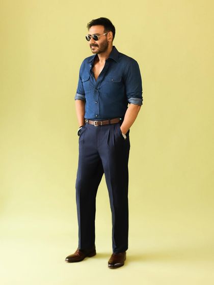 The full look, showing the dark wash denim shirt paired with navy pleated trousers and a stylish leather belt.