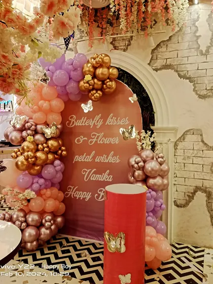A beautiful butterfly and flower-themed birthday decoration. The custom arched backdrop reads "Butterfly kisses & Flower petal wishes", surrounded by a delicate arrangement of peach, lavender, and gold balloons.
