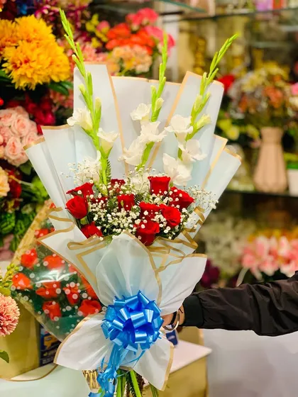 An elegant bouquet featuring red roses and tall white gladiolus flowers. The arrangement is wrapped in light blue and white paper with a striking blue bow.