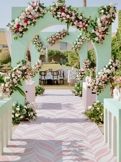 A fresh and elegant Mehendi entrance combining mint green arches with cascading arrangements of pink and white flowers. This pastel color palette creates a serene and sophisticated atmosphere for a daytime event.