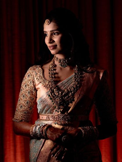 A dramatic portrait of the bride against a red curtain. The soft lighting highlights the intricate details of her blouse and jewelry.