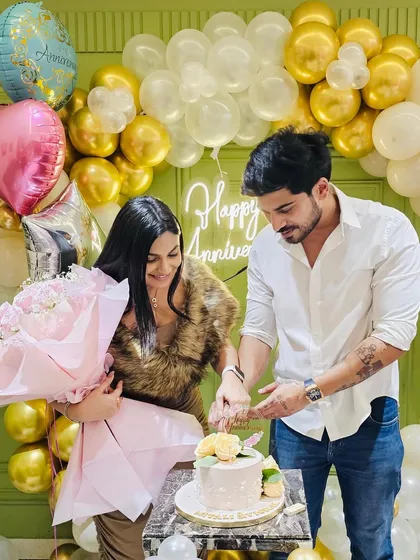 A couple cuts their cake in front of a gold and white balloon backdrop with a 'Happy Anniversary' neon sign. This setup is perfect for photos and celebrating together.