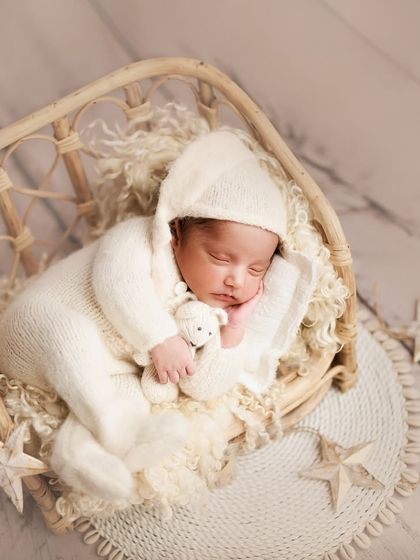 A dreamy, celestial-themed portrait. The baby is dressed in a white knit romper and bonnet, sleeping in a basket surrounded by little star props.