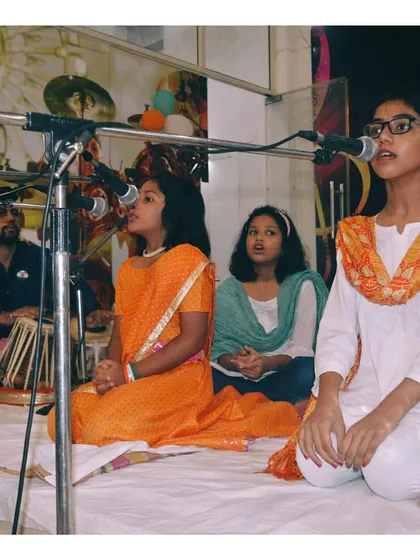 Students performing a group song in traditional attire, accompanied by a tabla player. Group singing helps develop skills in harmony and listening.