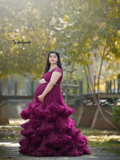 A regal portrait of a mother-to-be in a wine-colored ruffled gown and crown, set in a beautiful park.