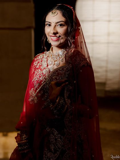 A stunning bride in a classic red lehenga, her smile lighting up the frame. The makeup is glamorous yet timeless, with a bold lip and defined eyes that make a statement.