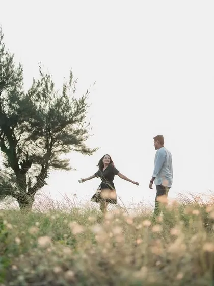 A wide shot capturing a couple in a field of wildflowers by the sea. The windswept tree and soft light create a dreamy, ethereal atmosphere for this pre-wedding portrait.