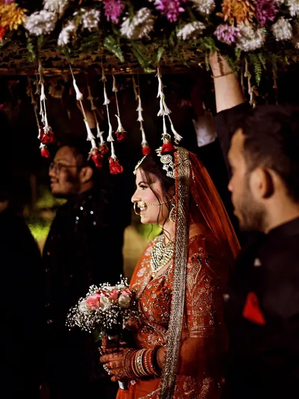 A side-profile shot of the bride entering under the phoolon ki chadar, holding a bouquet. The dark background makes her stand out beautifully.