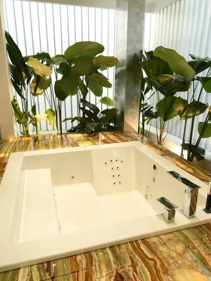 A luxurious jacuzzi tub set in a bed of green and gold onyx-style marble, surrounded by lush indoor plants. This design blurs the line between a bathroom and a tropical spa retreat.