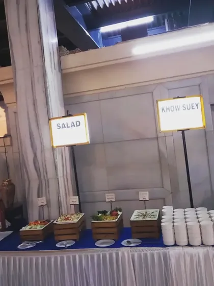 This is an example of a larger buffet setup for an event. I provide clear labeling for each station, like 'Salad' and 'Khow Suey', along with all the necessary plates and bowls, ensuring a smooth and organized dining experience for your guests.