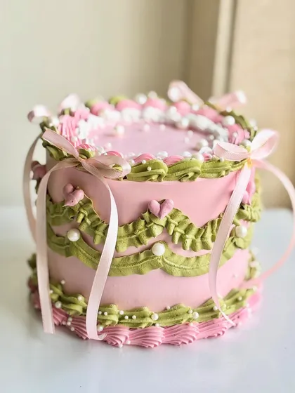 A dreamy vintage cake in pink and green, adorned with intricate piping, edible pearls, and delicate pink ribbons tied into bows for a fairytale finish.