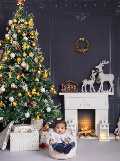 A tiny baby sits contently in a beanbag chair in front of a grand Christmas tree, looking like the most precious gift of all.