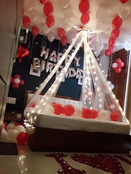 A cozy birthday room surprise with a canopy of fairy lights over the bed. Red and white balloons and rose petals on the floor add to the romantic and celebratory feel.