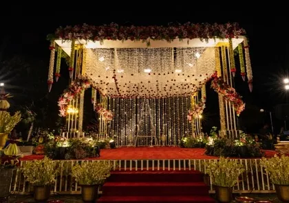A beautifully lit mandap at night, ready for a wedding ceremony. The combination of floral strings, a shimmering backdrop, and a red carpeted stage creates a dramatic and romantic setting.