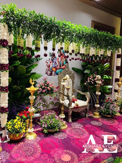 A complete Seemantham setup featuring a lush green backdrop with hanging floral garlands and a portrait of Krishna. The foreground is filled with beautifully arranged fruits and traditional offerings.