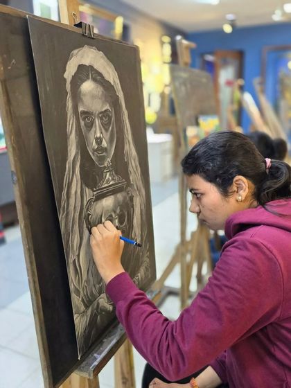 An artist at her easel, deeply focused on her large-scale charcoal drawing. The studio setting is visible in the background, filled with other creative works.
