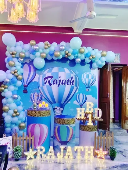A whimsical hot air balloon themed birthday party. The decor features a sky-blue backdrop with illustrated balloons, a custom balloon garland, and light-up marquee letters spelling the birthday boy's name.