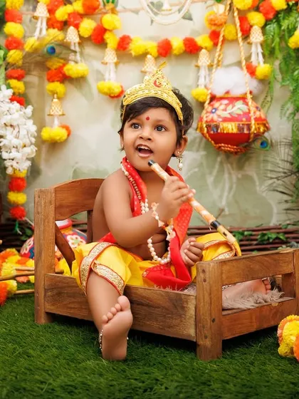 A baby Krishna sits on a miniature wooden bed, playing his flute in the festive Gokul setting.