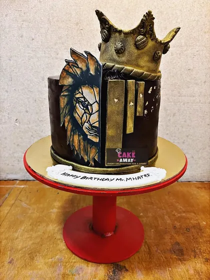 A powerful lion-themed cake with a crown. Half the cake is a rich chocolate finish, while the other half features a hand-painted edible image of a lion's face.
