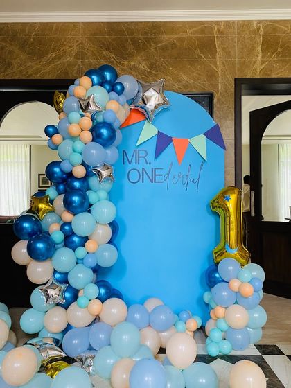A 'Mr. Onederful' first birthday party with a blue backdrop. The setup is decorated with a blue, peach, and white balloon garland and a number '1' foil balloon.