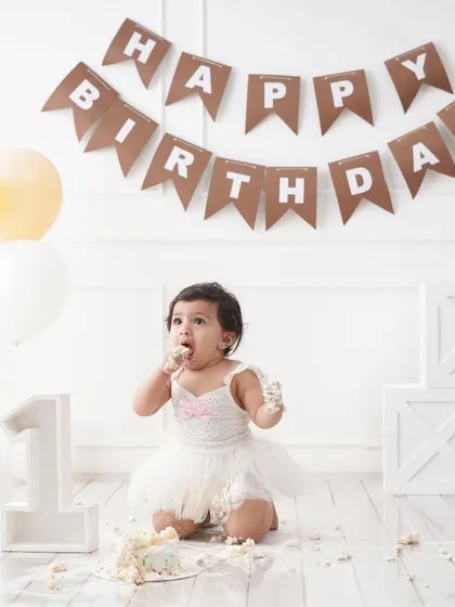 A fun shot of a baby girl enjoying her cake smash, with frosting on her face and a 'Happy Birthday' banner in the background.