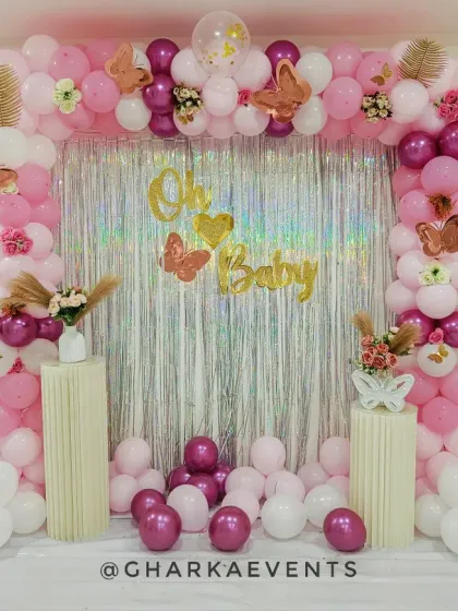 A glamorous pink and white baby shower setup. The silver tinsel backdrop is framed by a pink, white, and magenta balloon arch with butterfly accents.