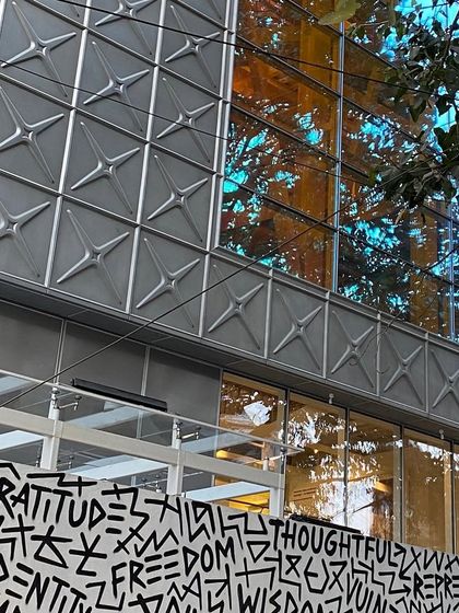 The reflection of trees on the dichroic glass of the MAP facade, with the graphic art wall below. This layering of art, architecture, and nature creates a complex and visually rich street presence.